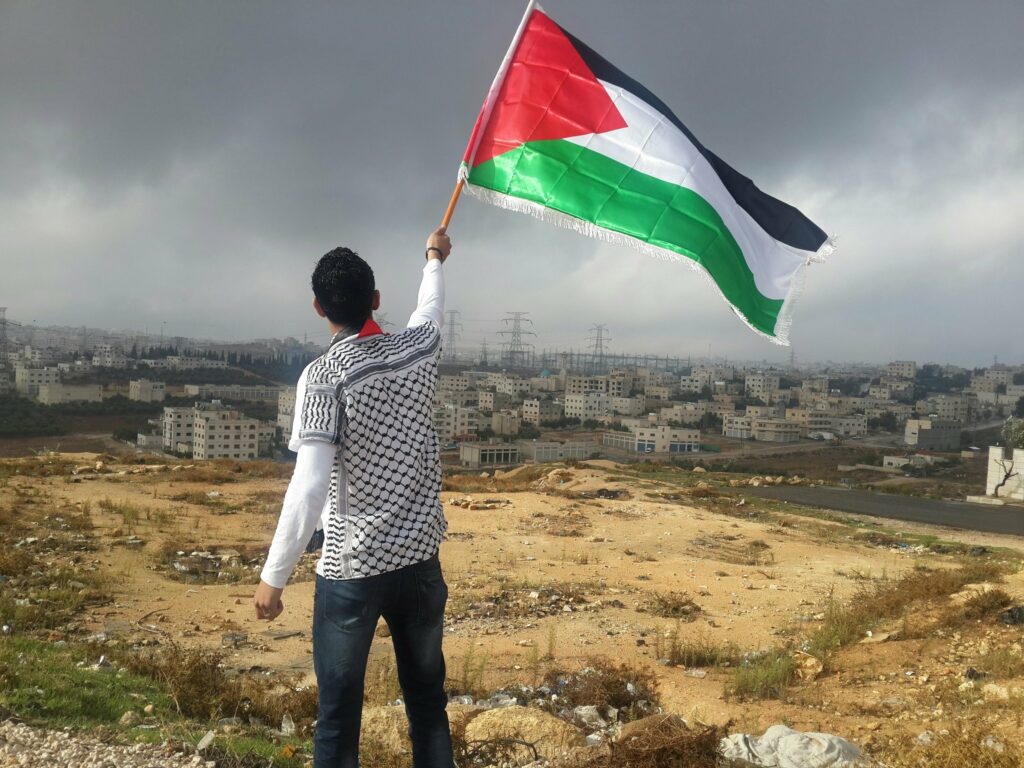 A young man waves a Palestinian flag out in a field facing a distant town as stormy clouds gather above.